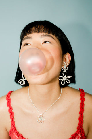 Woman blowing pink bubble gum bubble wearing floral earrings and silver Flora chain by Kara Yoo. 