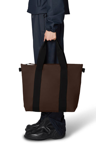 Woman holding RAINS tote bag in brown with black straps. 