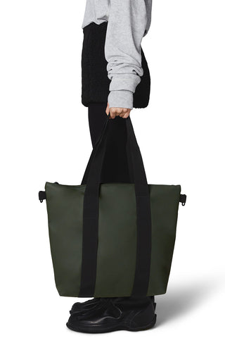 Woman holding RAINS tote bag in green with black straps. 