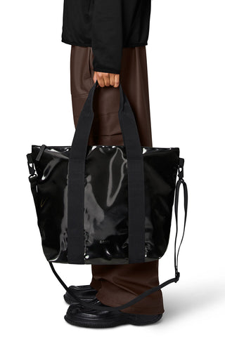 Model holding shiny black (Night) Tote Bag Mini by RAINS. 